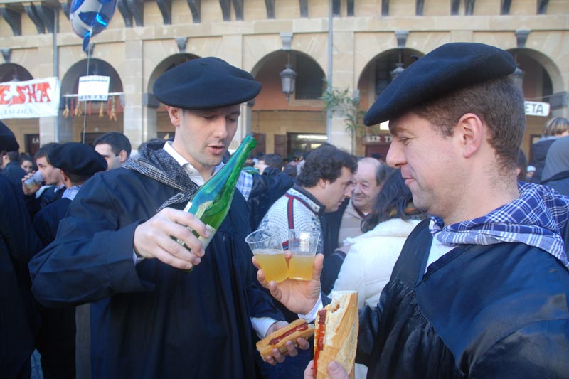 Personas vestidas de baserritarras bebiendo sidra y comiendo txistorra