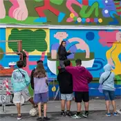 Varios jóvenes miran a una artista que hace un mural en la calle